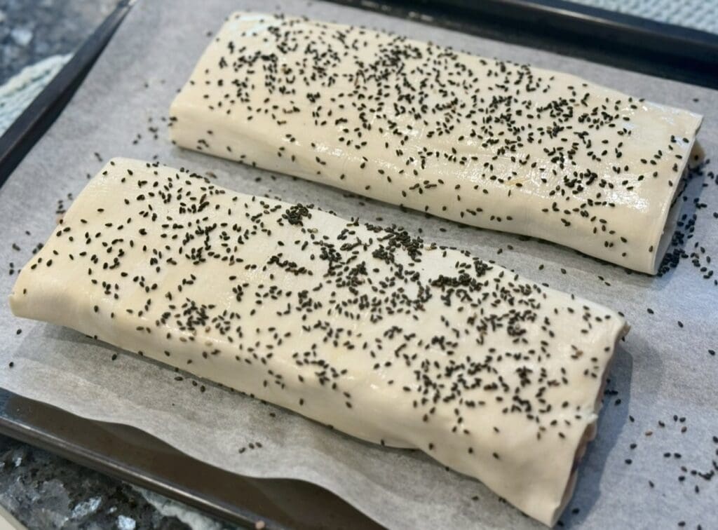 A tray holds two large sausage rolls, sprinkled with black sesame seeds, ready to bake.