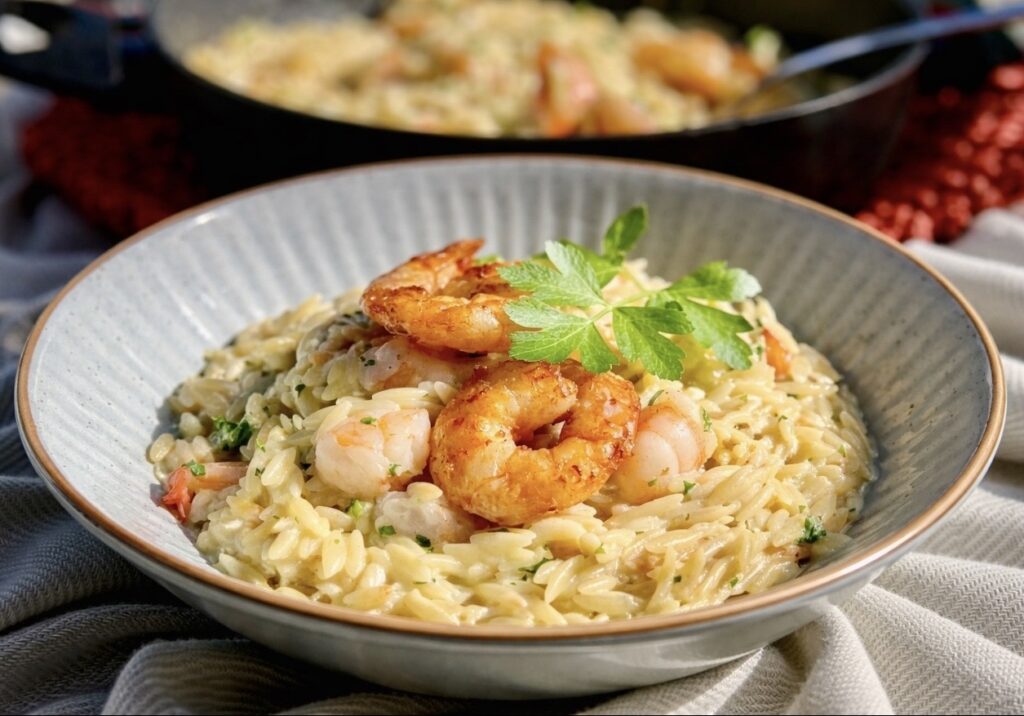 Creamy shrimp orzo served in a blue bowl topped with golden pan-seared shrimp and fresh parsley, with a skillet in the background