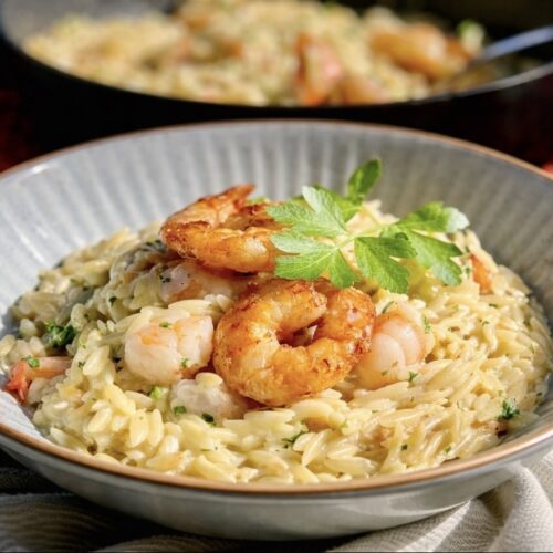 Creamy shrimp orzo served in a blue bowl topped with golden pan-seared shrimp and fresh parsley, with a skillet in the background