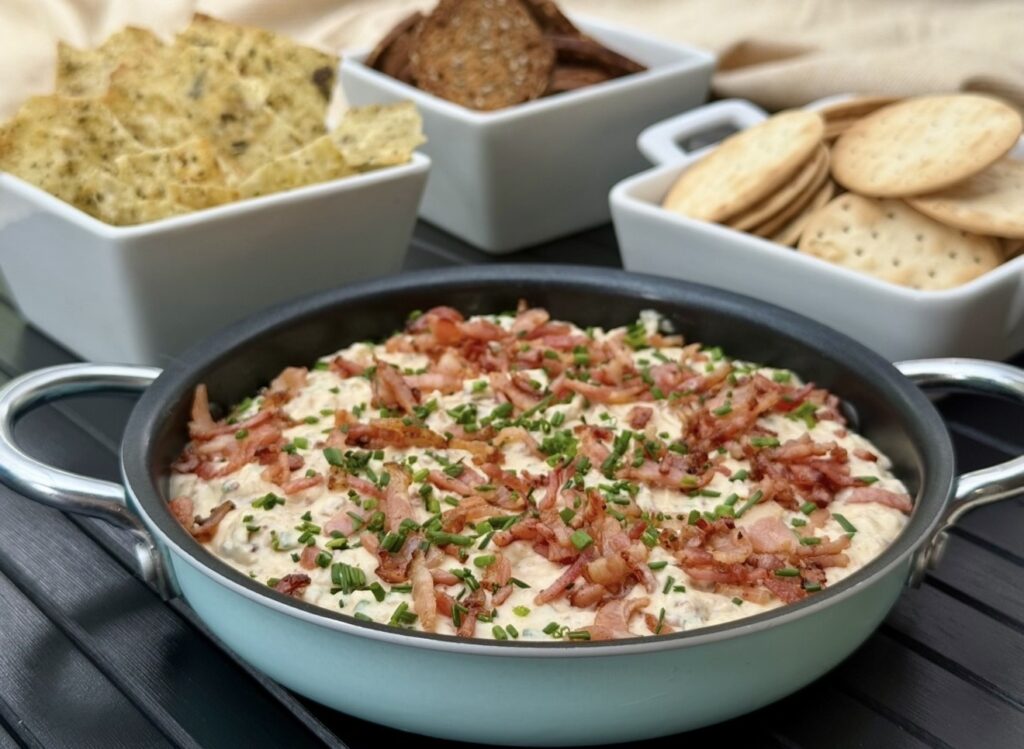 Creamy bacon and onion dip topped with crispy bacon and chives, served in a pan with crackers and chips for sharing