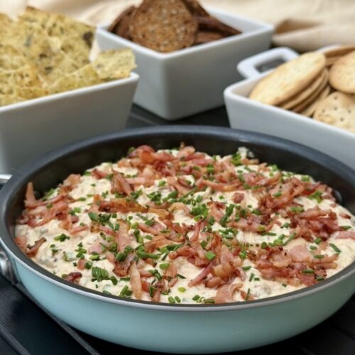 Creamy bacon and onion dip topped with crispy bacon and chives, served in a pan with crackers and chips for sharing