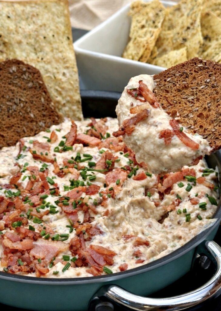 Close-up of creamy bacon and onion dip being scooped with a cracker, showing rich texture and crispy bacon pieces